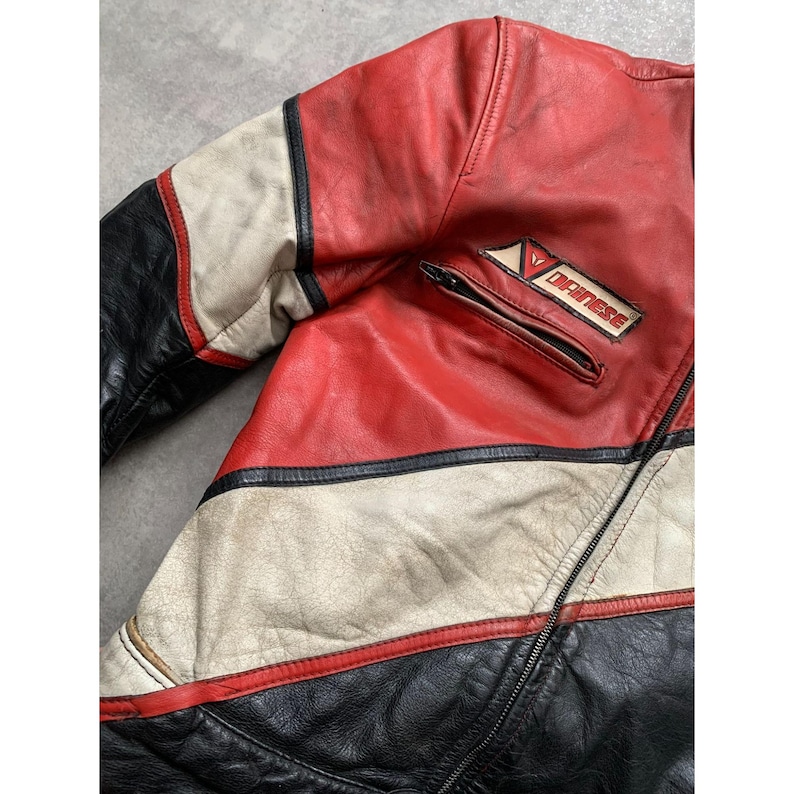 May include: A vintage red, black, and white leather motorcycle jacket. The jacket features a zippered pocket and a "DAINESE" patch. The jacket has a classic racing design with color-blocked panels and a worn appearance.