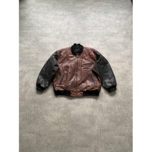 Könnte beinhalten: Braune und schwarze Leder-Bomberjacke mit Reißverschluss. Die Jacke hat schwarze Ärmel und einen braunen Körper. Kragen, Manschetten und Saum sind schwarz. Eine kleine Tasche befindet sich auf der linken Brust.