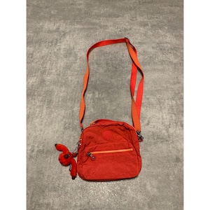 Op de afbeelding: Een rode crossbodytas met een verstelbare riem die overgaat van rood naar oranje. De tas heeft een hoofdvak met ritssluiting en een kleine, bevestigde rode apen sleutelhanger. De tas is gemaakt van een duurzaam, getextureerd materiaal.