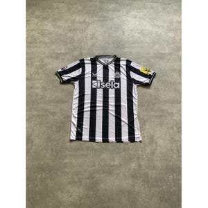 May include: A short-sleeved Newcastle United soccer jersey featuring vertical black and white stripes, a V-neck collar, and the text sela printed in white across the chest. The shirt includes a team crest, a brand logo, and sleeve patches.