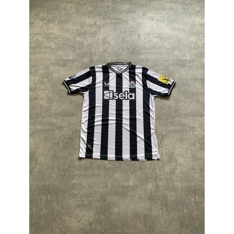 May include: A short-sleeved Newcastle United soccer jersey featuring vertical black and white stripes, a V-neck collar, and the text sela printed in white across the chest. The shirt includes a team crest, a brand logo, and sleeve patches.