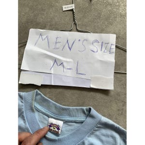 May include: A light blue t-shirt with a ribbed crew neckline and a white interior tag featuring the Rip Curl logo. A white paper sign is attached to a metal hanger above the shirt, with the handwritten text MEN'S SIZE M-L in blue ink.