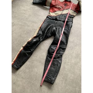 May include: A black, red, and cream-colored leather motorcycle racing suit. The suit has a full-length zipper, padded knees, and red and cream stripes down the sides. A pink measuring tape is laid across the suit.