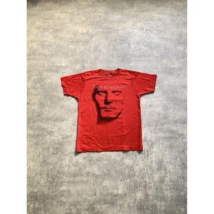 Peut inclure: Un t-shirt rouge vif &agrave; manches courtes avec une impression graphique noire repr&eacute;sentant un visage humain au centre. Au-dessus du visage, le texte "MADE IN GERMANY" est imprim&eacute; dans une police simple et claire. Le t-shirt a un col rond.