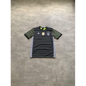 Könnte beinhalten: Ein kurzärmeliges Sporttrikot mit dunkelgrau horizontal gestreiftem Rumpf und olivgrünen Ärmeln. Das Trikot zeigt ein weißes Adidas-Logo, ein goldenes Emblem und ein kreisförmiges Mannschaftswappen mit vier Sternen auf der Brust sowie weiße Seitenstreifen.