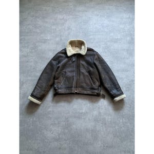 May include: A brown leather jacket with a cream-colored shearling collar and cuffs. The jacket has a zipper closure and a belt at the waist. The jacket is a classic style, perfect for cold weather.