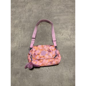 Op de afbeelding: Roze schoudertas met bloemenprint, bijpassende riem en een kleine, paarse apen sleutelhanger. De tas heeft een klepsluiting en een klein vak met ritssluiting. De tas is ongeveer 25 cm breed.
