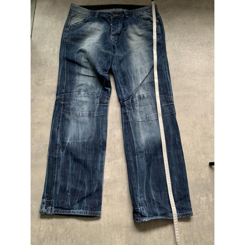 May include: Blue denim jeans with a faded wash and a relaxed fit. The jeans feature a button fly, multiple pockets, and reinforced knee panels. A measuring tape is placed along the side of the jeans.