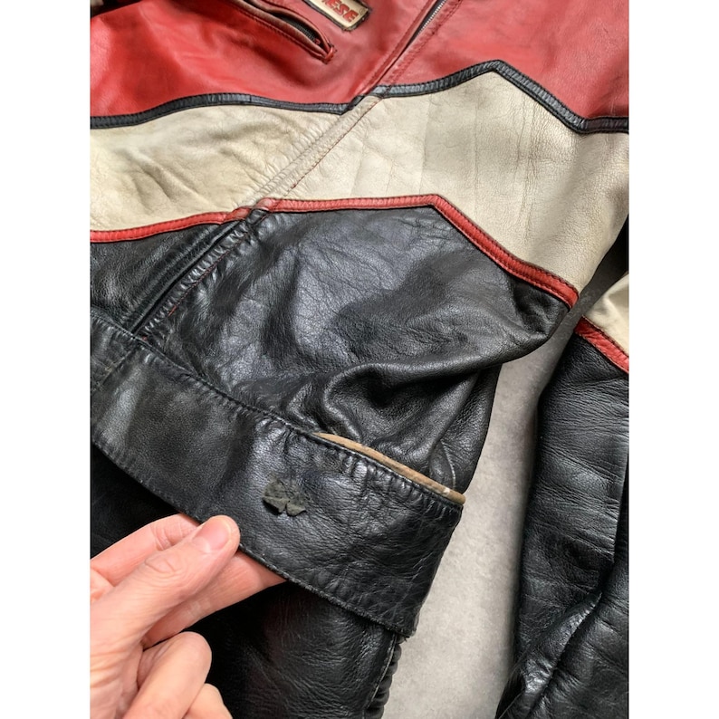 May include: Close-up of a vintage leather jacket with a color block design. The jacket features red, white, and black panels with red trim. The leather appears worn, adding to its vintage appeal. A small emblem is visible on the cuff.