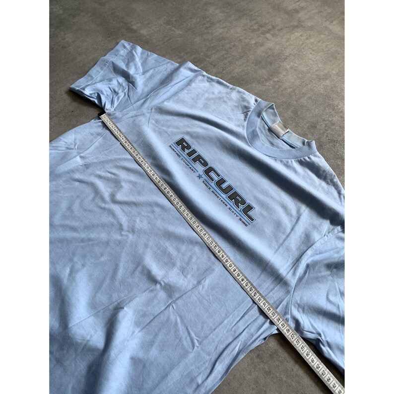 May include: A light blue short-sleeve t-shirt featuring the text RIPCURL SURFING COMPANY SINCE NINETEEN SIXTY NINE in black and white lettering across the chest. A white measuring tape is placed diagonally across the shirt for size reference.