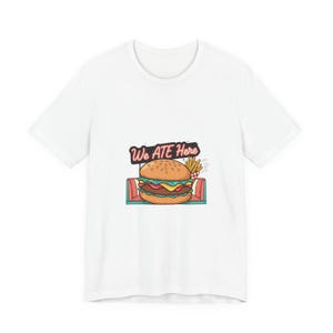 May include: White t-shirt featuring a colorful graphic of a hamburger with fries and the text "We Ate Here" in a retro style. The burger has a sesame seed bun, a patty, cheese, and other toppings.