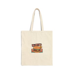 May include: Cream-colored tote bag with a retro-style burger and fries graphic. The graphic includes the text "We Ate Here" in a vintage font. The bag has a simple design with two handles.