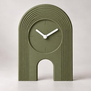 May include: An olive green table clock with an arched silhouette and concentric grooved lines. The clock face is circular with simple white hour and minute hands, lacking numerals, and features a small arched cutout at the base.
