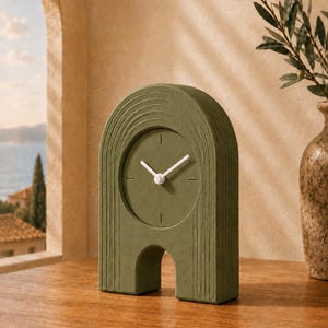 May include: An olive green arch-shaped table clock with a textured, ribbed surface. The clock face is circular and matches the body colour, featuring simple white hour and minute hands. The design includes a central cutout at the base.