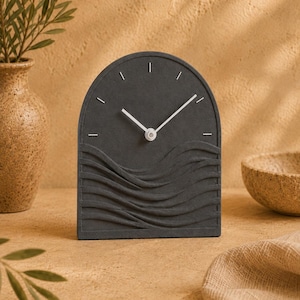 May include: An arched tabletop clock in a dark charcoal gray finish. The clock face features a textured, wave-like relief pattern across the bottom half, complemented by simple white hour markers and slender white clock hands at the center.