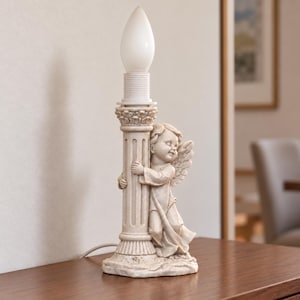 May include: A table lamp featuring a cream-colored stone-like base sculpted as a cherub with wings embracing a fluted column. The top of the column holds a white, flame-shaped light bulb in a threaded socket, creating a vertical structure.