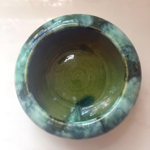 May include: A round ceramic bowl featuring a glossy finish with a deep olive green interior and a mottled exterior pattern of blended teal, dark blue, and sage green tones. The thick, rounded rim transitions into a smooth, concave basin.
