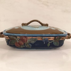 May include: Oval ceramic casserole dish with a lid. The dish is navy blue with a woven basket design and features painted fruit and leaves. The lid is brown and light blue with a brown handle. The dish measures approximately 12 inches long.