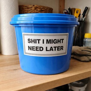 Pode incluir: Um balde de plástico azul brilhante com um rótulo branco que diz "SHIT I MIGHT NEED LATER" em texto preto. O balde está em uma prateleira de madeira, com outros itens ao fundo.