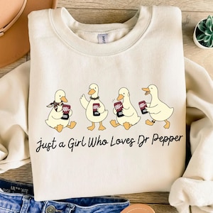 May include: Cream-colored sweatshirt featuring four cartoon ducks holding Dr Pepper cans. The ducks are in various poses, and the text "Just a Girl Who Loves Dr Pepper" is printed below the ducks.