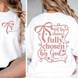 May include: White sweatshirt with a mauve bow and text. The front of the sweatshirt has a bow graphic and the text "Romans 4:7-8". The back of the sweatshirt has the text "not by chance, fully chosen by God, Romans 4:7-8".