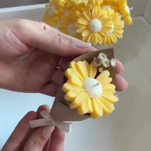 Peut inclure: Un petit bouquet de bougies en forme de marguerites jaunes enveloppé dans du papier brun et noué avec un ruban blanc. Le bouquet présente une marguerite jaune proéminente avec un centre blanc, agrémentée de petites fleurs blanches séchées.