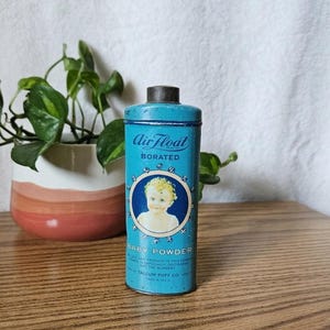 Könnte beinhalten: Vintage Baby-Puder-Dose aus tealfarbenem Metall mit schwarzem Deckel. Die Dose trägt die Aufschrift "Air Float Borated" und ein rundes Bild eines Babys. Die Worte "Baby Powder" sind darunter aufgedruckt. Die Dose steht auf einer Holzoberfläche.
