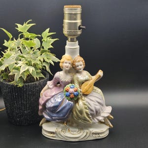 May include: A porcelain lamp base featuring two figures sitting together on an oval pedestal. One figure wears a lavender and blue dress holding a bouquet of flowers, while the other wears a green and lavender dress and holds a lute instrument.