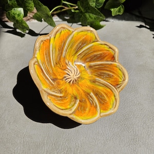 May include: A ceramic bowl shaped like a flower with swirling petals in vibrant shades of yellow and orange. The centre features a textured, raised design, and the edges of the petals are defined by a smooth, light tan glazed rim.