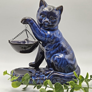 May include: A glossy dark blue ceramic cat figurine with a mottled finish. The cat is in a seated position, holding a small, suspended black metal bowl with a chain in one raised paw. A circular base is attached to the cat's feet.