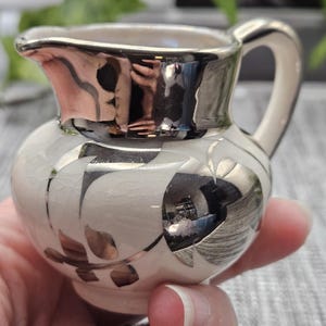 Könnte beinhalten: Ein kleines Sahnekännchen mit cremefarbener Basis und einer Craquelé-Glasur. Das Kännchen ist mit metallischen Silberblattmustern verziert und verfügt über einen reflektierenden silbernen Rand sowie einen silbernen Henkel.