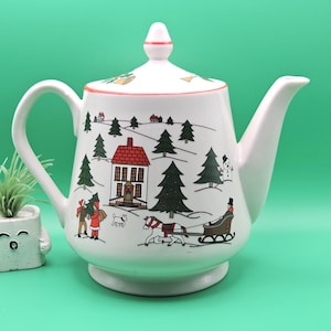 May include: A white ceramic teapot with a lid featuring a festive winter scene. The illustration includes a red-roofed house, evergreen trees, a horse-drawn sleigh, a snowman, and figures in winter clothing. The teapot has a curved handle and spout.