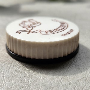 May include: A round, off-white, fluted disc with a black base. The top surface features a brown line-art illustration of a flower and a banner containing the text "PRINCESS" and "CHICAGO" printed in a clear, dark brown, sans-serif font.