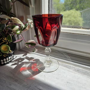 Puede incluir: Una copa de cristal de color rojo intenso con un cuenco ancho y redondeado que presenta hendiduras verticales. El tallo es de cristal transparente y facetado, y termina en una base circular. El cristal refleja la luz y proyecta una sombra roja.