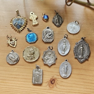 Peut inclure: Une collection de quatorze pendentifs et breloques religieux assortis de formes et tailles variées, avec des finitions en métal argenté et doré. Les pièces incluent des représentations de mains en prière, de saints et de cœurs sacrés émaillés bleus.