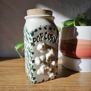 May include: A cream-colored ceramic canister with a cork lid, featuring the word POPCORN in black lettering and raised, three-dimensional white popcorn shapes on the front. The sides are decorated with a green speckled pattern.