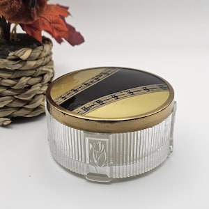 May include: A round glass powder jar with a clear, vertically ribbed body featuring embossed tulip motifs. The lid is metallic gold with a central black diagonal band decorated with intricate gold geometric patterns and a gold-toned rim.