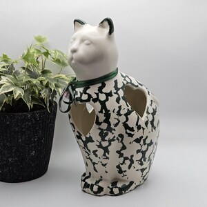 May include: A ceramic cat figurine with a white base and a black speckled pattern. The cat has a smooth white head, black-tipped ears, and a dark green ribbon tied around its neck. It features two heart-shaped cutouts on its body.