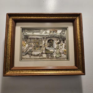 May include: Framed artwork depicting a detailed, grayscale scene of a shop with figures and a baby carriage. The artwork is set within a gold-toned wooden frame, adding a classic touch. The image captures a quaint, old-fashioned setting, perfect for home decor.