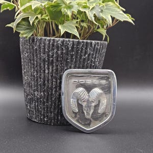 Puede incluir: Un emblema de vehículo Dodge de color plateado que presenta un diseño de cabeza de carnero en relieve centrado dentro de un marco en forma de escudo. La palabra DODGE está grabada en letras mayúsculas en la parte superior de la insignia metálica, que tiene un acabado pulido.