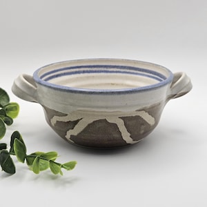 May include: A ceramic bowl with two side handles, featuring a white interior with blue horizontal stripes near the rim. The exterior has a dark brown base with a white, jagged, mountain-like pattern encircling the middle section of the bowl.