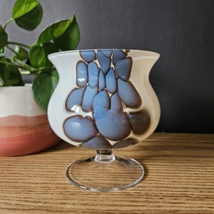 May include: A decorative glass vase with a wide, curved top and a clear, circular base. The vase features a white background with blue and brown abstract shapes. The vase is on a wooden surface, with a potted plant in the background.