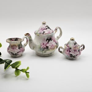 Puede incluir: Un juego de té en miniatura de porcelana que incluye una tetera, una jarra de leche y un azucarero con tapa. Cada pieza está decorada con un delicado patrón floral rosa y detalles en oro en las asas, boquillas y tapas.