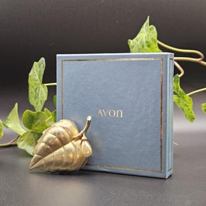 Op de afbeelding: Een goudkleurige metalen bladbroche met een crèmekleurig druppelvormig parelaccent. De broche is geplaatst voor een vierkante, lichtblauwe geschenkdoos met een gouden rand en het woord AVON in gouden letters op de voorzijde gedrukt.