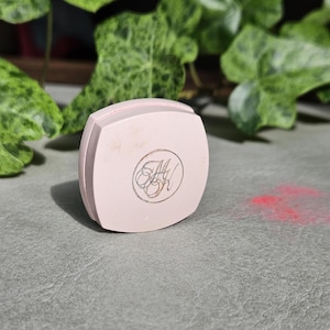 May include: A square, light pink cosmetic compact case with rounded corners. The lid features a circular emblem with the stylized letters "MK" printed in a dark, thin font. The compact has a smooth, matte finish and is positioned upright.