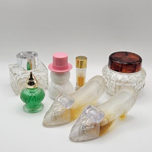 May include: A collection of decorative glass items including two frosted glass shoes with bow details, a green glass bottle with a gold cap, a snowman-shaped bottle with a pink hat, a square glass bottle, a tall thin bottle, and a glass jar with a brown lid.