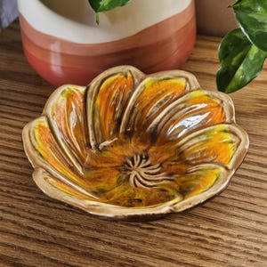 Könnte beinhalten: Eine blütenförmige Keramikschale mit einem leuchtenden orange-gelben Blumenmuster. Die Schale hat einen braunen Rand und steht auf einer Holzoberfläche. Die Schale ist wahrscheinlich vintage und für dekorative Zwecke geeignet.
