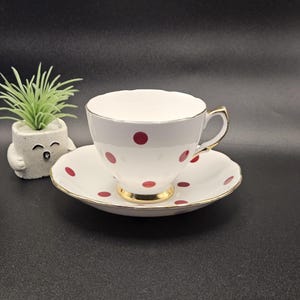 May include: A white porcelain teacup and saucer set featuring a pattern of small red polka dots. The teacup has a gold-trimmed handle and rim, and the saucer also features a gold-trimmed edge. A small, light green artificial plant sits nearby.