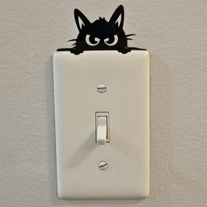 Può includere: Una copertura per interruttore a forma di gatto nero sbircia sopra una placca interruttore bianca. Il gatto ha grandi occhi espressivi e orecchie a punta. L'interruttore è in posizione 'on'. Questo oggetto decorativo aggiunge un tocco giocoso a qualsiasi stanza.