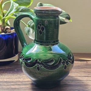Op de afbeelding: Een groene keramische kan met een handvat en een afgeronde body. De kan heeft donkere accenten en een decoratief patroon rond de basis. De rand is bruin. De kan staat op een houten oppervlak.